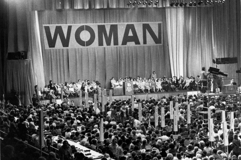 Black and white photo of a large crowd gathered at an indoor event with a stage backdrop displaying the word WOMAN